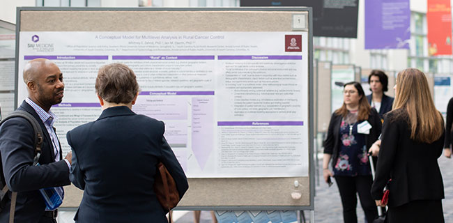 Photo of a two men looking at a poster presentation at a conference, with other people in the background. 